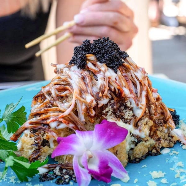 A large, ornate sushi roll topped with black caviar, sauces, and a purple orchid on a blue plate.