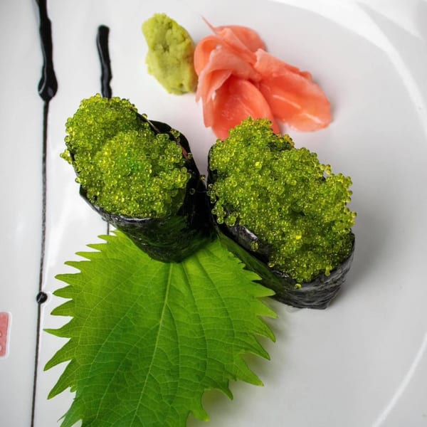 Two pieces of green tobiko gunkan sushi with wasabi, ginger, and a shiso leaf on a white plate.