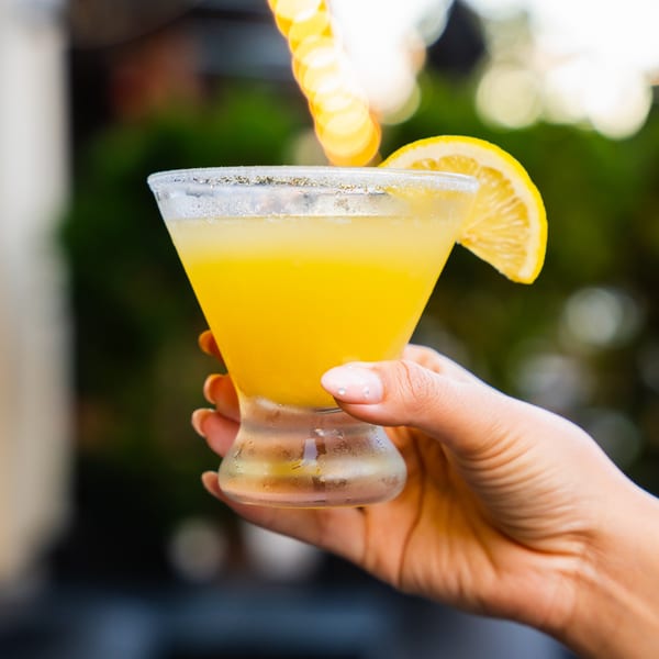 A hand holds a vibrant yellow cocktail in a martini glass, garnished with a lemon slice.