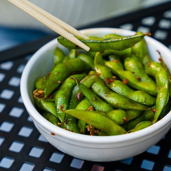 Chopsticks picking up a single edamame pod from a white bowl filled with seasoned edamame.