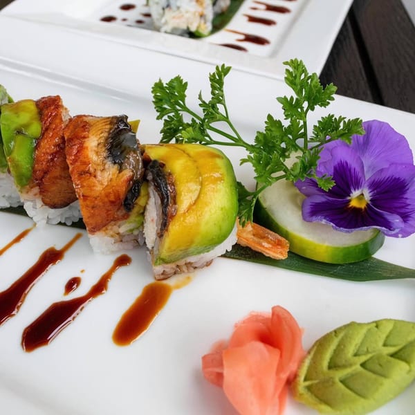 Sinju Sushi's Dragon Roll, featuring eel and avocado, garnished with parsley, a purple flower, and sauce.